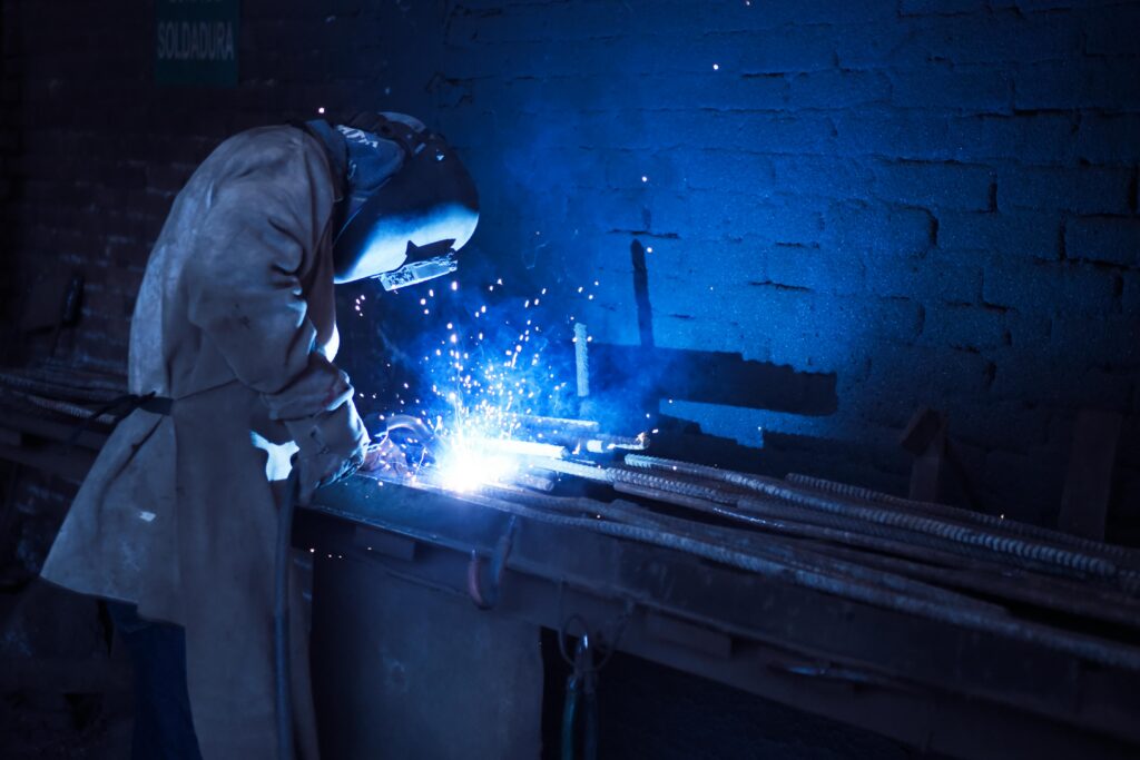 pexels photo 1474993 1474993 Industrial worker welding metal indoors, emitting bright blue sparks and smoke.